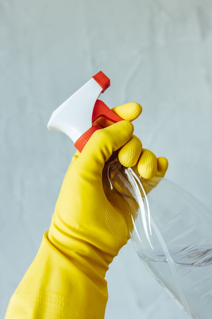 hero-img-02 A hand wearing a yellow rubber glove holds a red and white spray bottle against a neutral background.